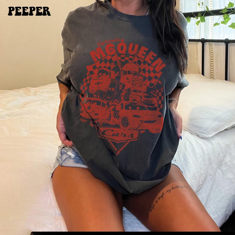 May include: Dark gray t-shirt featuring a red graphic of race cars and the text "McQueen Cars". The shirt has short sleeves and a crew neck. The word "PEEPER" is printed in the upper left corner.
