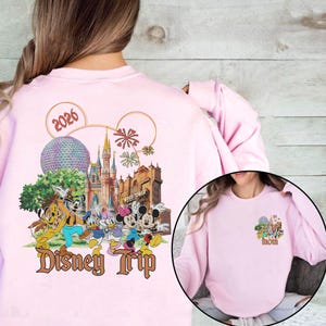 May include: Light pink sweatshirt featuring a colorful Disney Trip design with Mickey Mouse, Minnie Mouse, Donald Duck, and other characters. The design includes the text "Disney Trip" and the year "2026". A smaller design is on the front.