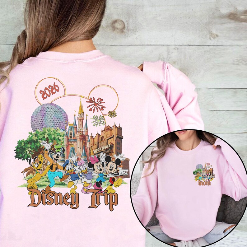 May include: Light pink sweatshirt featuring a colorful Disney Trip design with Mickey Mouse, Minnie Mouse, Donald Duck, and other characters. The design includes the text "Disney Trip" and the year "2026". A smaller design is on the front.