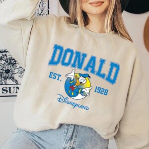 May include: Beige sweatshirt featuring the name "DONALD" in blue block letters, with "EST. 1928" and a graphic of Donald Duck. The word "Disneyland" is printed below the graphic. The sweatshirt has a crew neck.