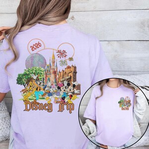 May include: A light purple t-shirt featuring a colorful graphic of Disney characters and landmarks. The design includes the text "Disney Trip" and the year "2026". A smaller graphic on the front says "Mom".