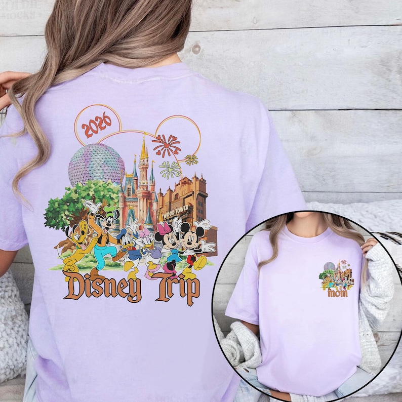 May include: A light purple t-shirt featuring a colorful graphic of Disney characters and landmarks. The design includes the text "Disney Trip" and the year "2026". A smaller graphic on the front says "Mom".