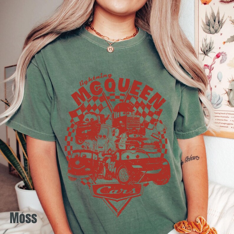 May include: Moss green t-shirt featuring a red graphic of Lightning McQueen and other characters from the Cars movie. The design includes the text "Lightning McQueen" and "Cars" with a checkered flag background. The shirt is a crew neck.