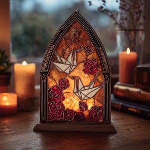 Op de afbeelding: Een gebrandschilderde lamp in een gotische boogvorm. Het heeft witte origami-kraanvogels en rode rozen op een amberkleurige achtergrond. De lamp, op een houten voet, wordt van binnenuit verlicht en geeft een warm licht.