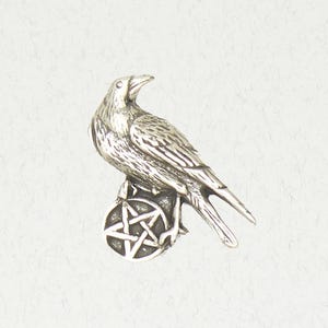 May include: A silver-colored raven figurine perched atop a circular base with a five-pointed star design. The raven is detailed with textured feathers and a curved beak. The figurine is likely a decorative item or a piece of jewelry.