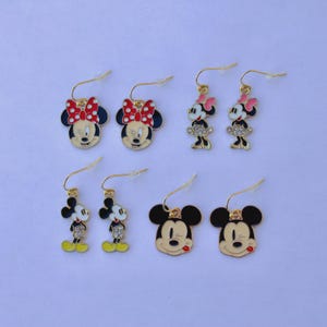 May include: A collection of gold-toned earrings featuring iconic cartoon characters. Designs include Minnie Mouse with red polka dot bows and Mickey Mouse with a red tongue. Some earrings have rhinestone accents.