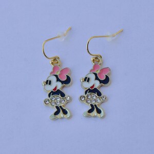 May include: A pair of gold-toned dangle earrings featuring a cartoon character design. Each earring showcases a character with a pink bow, black dress, and a skirt adorned with clear rhinestones. The earrings have gold-colored hooks.