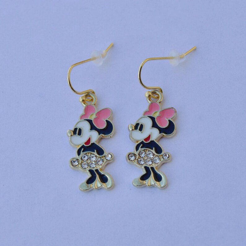 May include: A pair of gold-toned dangle earrings featuring a cartoon character design. Each earring showcases a character with a pink bow, black dress, and a skirt adorned with clear rhinestones. The earrings have gold-colored hooks.