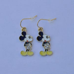 May include: A pair of gold-tone Mickey Mouse dangle earrings. Each earring features a black head, white face, red shorts, and yellow shoes. The earrings are designed with a hook closure.