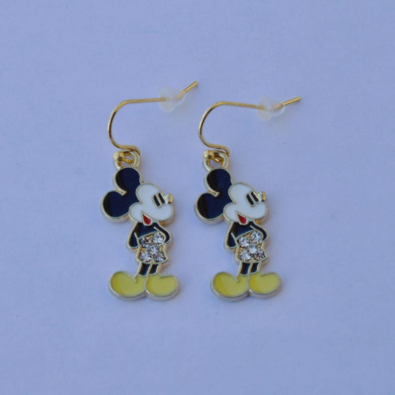 May include: A pair of gold-tone Mickey Mouse dangle earrings. Each earring features a black head, white face, red shorts, and yellow shoes. The earrings are designed with a hook closure.