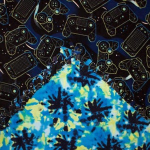 May include: A two-sided fleece blanket. One side features a pattern of neon green and black video game controllers on a dark blue background. The other side has a tie-dye design in shades of blue and yellow. The edges are knotted.