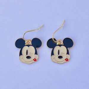 May include: A pair of gold-toned dangle earrings featuring the face of a cartoon mouse with black ears, a cream-colored face, and a red tongue. The earrings have a gold-colored hook.