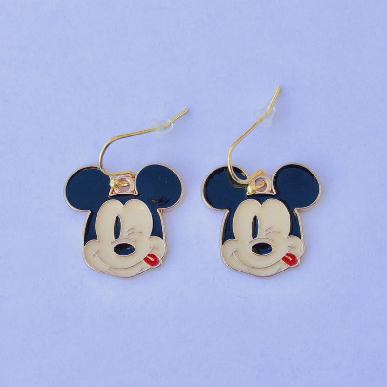 May include: A pair of gold-toned dangle earrings featuring the face of a cartoon mouse with black ears, a cream-colored face, and a red tongue. The earrings have a gold-colored hook.