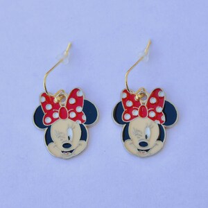May include: A pair of Minnie Mouse earrings. Each earring features a black and cream-colored face with a red bow with white polka dots. The earrings have gold-colored hooks.