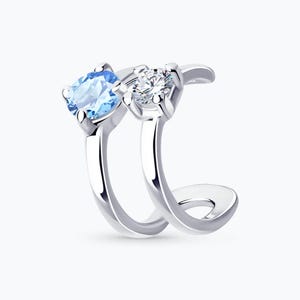 May include: A silver-toned ear cuff featuring two gemstones. One is a light blue oval-cut stone, and the other is a clear, round-cut stone. The cuff curves around the ear, creating a modern and elegant look. Jewelry.