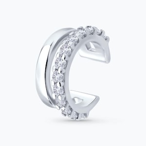 May include: A silver ear cuff with a double-band design. One band is smooth and polished, while the other is adorned with a row of small, clear gemstones. The cuff curves around the ear, creating a modern and elegant look.
