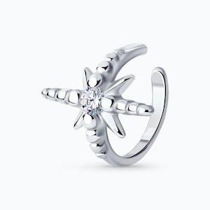 May include: A silver ear cuff featuring a starburst design with a central clear gemstone. The cuff wraps around the ear, with a decorative beaded crescent shape and a star-shaped design with pointed edges. The jewelry is set against a white background.