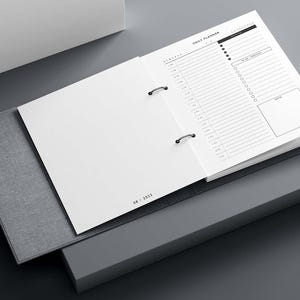 Può includere: Un'agenda giornaliera aperta, con una copertina grigia e un design minimalista. Le pagine bianche presentano un programma e una lista di cose da fare in nero. La data "08 / 2025" è stampata in basso a sinistra.