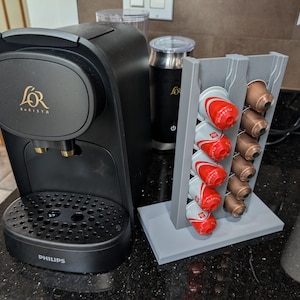 以下が含まれることがあります： 金色のアクセントが付いた黒いPhilips L'OR Baristaコーヒーマシンとポッドホルダー。グレーのポッドホルダーには、赤と茶色のコーヒーポッドが入っています。黒いミルク泡立て器も表示されています。