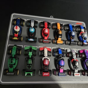 May include: A collection of miniature race cars in various colors, including blue, red, green, and orange, arranged on a gray display board. Each car features detailed designs and sponsor logos, showcasing a Formula 1 theme.
