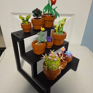 May include: A black, three-tiered display stand holds a collection of miniature potted plants. The plants include red flowers, green cacti, and other colorful blooms. The pots are brown and the plants are made of plastic.