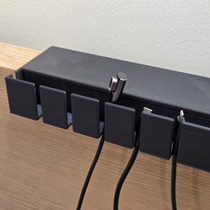 May include: A black cable organizer with multiple slots, mounted on a wooden surface. Several black cables are inserted into the slots. A silver-colored charging cable is also visible.