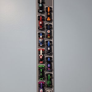 May include: A vertical wall-mounted display rack featuring a gray racetrack design with a checkered finish line at the top. The rack holds fourteen miniature Formula 1 style race cars in various colors including black, orange, red, blue, green, and purple.