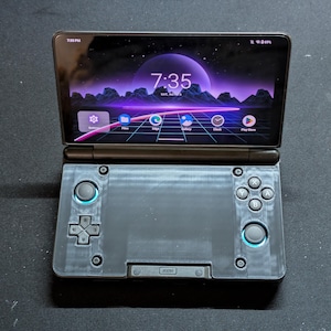 May include: A handheld gaming device with a black screen displaying a purple and blue landscape with the time 7:35 PM. The device has a gray body with a D-pad, buttons, and joysticks with blue lights.