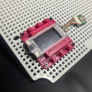 May include: A small, pink electronic device with a black screen and a clear plastic cover. It is mounted on a white grid and connected to a circuit board with colorful wires. The device has a metallic component on the side.