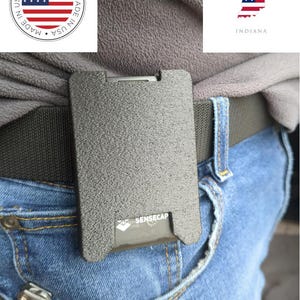 May include: A textured, gray Sensecap wallet clipped to a dark green belt, worn with blue jeans. The wallet is rectangular with a small logo and text. A "Made in USA" emblem is in the upper left corner, and the Indiana state outline with the American flag is in the upper right.