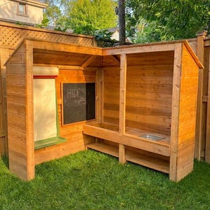 May include: A wooden playhouse with a chalkboard and a roll-up paper dispenser. The playhouse is made of natural wood and has a small sink. The words "Hide + play" are written on the chalkboard.