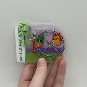 Peut inclure: Un porte-clés en acrylique transparent avec des personnages de dessins animés et le texte "BATTLE FOR BEDI". Le porte-clés a un clip en fil de fer vert et un design coloré avec une roue de Ferris et d'autres éléments animés. Le porte-clés est tenu par une main.