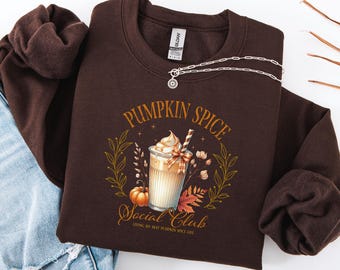 May include: A brown sweatshirt with the text "Pumpkin Spice Social Club" and a graphic of a pumpkin spice latte. The design includes a pumpkin, leaves, and floral elements. A silver necklace is draped across the sweatshirt.