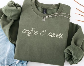 May include: An olive green sweatshirt with the words "coffee & books" in white script. A silver necklace with a circular pendant rests on the sweatshirt. The sweatshirt is folded, with a portion of blue jeans visible.