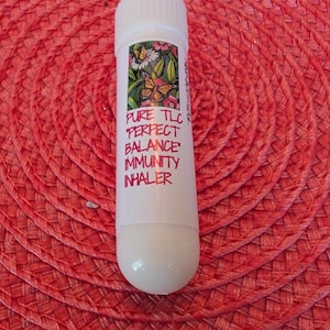 May include: A white inhaler with a white cap and a rounded bottom. The inhaler has a label with the words "PURE TLC PERFECT BALANCE IMMUNITY INHALER" and a floral and butterfly design. The inhaler is on a red woven surface.