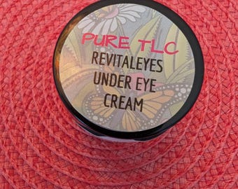 May include: A small, round jar of Pure TLC Revitaleyes Under Eye Cream. The label features the product name in pink and white text, with a floral and butterfly design. The jar is set against a red woven background.