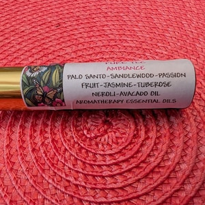 May include: A gold-topped aromatherapy essential oil roller bottle. The label displays a butterfly and floral design, with ingredients including Palo Santo, Sandalwood, Passion, Fruit, Jasmine, Tuberose, Neroli, and Avocado Oil. The bottle rests on a red surface.