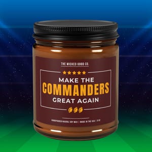 May include: A brown glass jar candle with a black lid. The label reads "MAKE THE COMMANDERS GREAT AGAIN" in gold lettering. The candle is hand-poured natural soy wax and made in the USA.