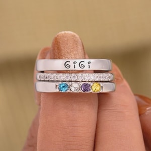 May include: Three silver rings stacked together. The top ring is engraved with the word "Gigi". The middle ring is encrusted with small clear stones. The bottom ring has four coloured gemstones: blue, purple, and yellow.