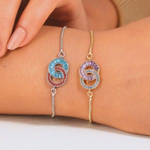 Puede incluir: Dos pulseras con círculos entrelazados, una plateada y otra dorada. La pulsera plateada presenta un círculo con gemas azules y rojas, mientras que la pulsera dorada tiene gemas moradas y azules. Ambas están en una muñeca.