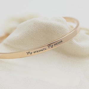 May include: A gold-toned metal bangle bracelet with a smooth, polished finish. The outer surface of the bracelet is engraved with the black text "My STRENGTH. My ANCHOR" along with two small anchor symbols placed at the beginning and end of the phrase.