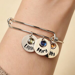 May include: Silver bangle bracelet featuring three circular charms. Each charm has a gemstone and a name: Lisa, Mary, and Lily. The gemstones are green, blue, and orange. The bracelet is worn on a wrist.