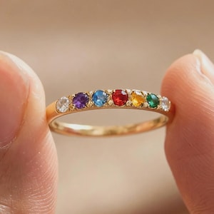 May include: A gold ring with a series of colourful gemstones arranged in a rainbow sequence. The gemstones include white, purple, blue, red, yellow, and green. The ring is held between two fingers against a blurred background.