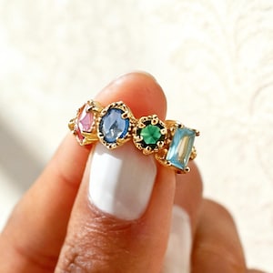 Peut inclure: Une bague en or ornée de cinq pierres précieuses colorées : rose, bleu, vert et turquoise. Les pierres sont serties dans une monture dorée décorative. La bague est tenue par une main avec du vernis à ongles blanc.