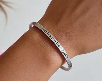 May include: A silver-toned metal cuff bracelet featuring the engraved text "besties ♡ tribe ♡ family" in a clean, sans-serif font. The bracelet has a smooth, polished finish and a simple, open-ended design that wraps around the wrist.