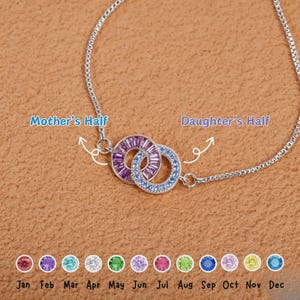Peut inclure: Bracelet en argent avec deux cercles entrelacés, un violet et un bleu, ornés de pierres précieuses. Le texte indique "Mother's Half" et "Daughter's Half". Des pierres précieuses colorées représentent les mois de janvier à décembre.