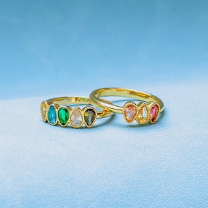 Könnte beinhalten: Zwei goldfarbene Ringe mit tropfenförmigen Edelsteinen in verschiedenen Farben, darunter Blau, Grün und Rosa. Die Ringe sind vor einem hellblauen Hintergrund platziert, der die lebendigen Farben und das Design des Schmucks hervorhebt.