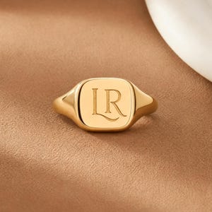 Puede incluir: Un anillo de sello dorado con una superficie lisa y pulida que presenta las letras LR grabadas en el centro de la cara cuadrada. El anillo tiene una banda sólida que se estrecha hacia la parte posterior, con un acabado metálico definido.