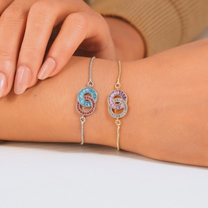 May include: Two bracelets with interlocking circles, one silver and one gold. The silver bracelet features a light blue and red gemstone design. The gold bracelet has a purple and light blue gemstone design. Both bracelets are on a wrist.