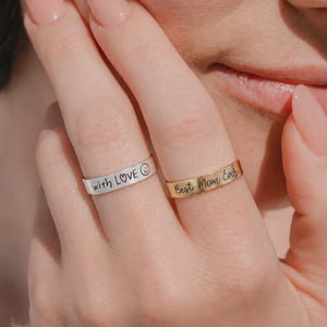 以下が含まれることがあります： 隣り合う指にはめられた2つの平らなバンドリング。1つはシルバーカラーで「with LOVE」という文字とスマイリーフェイスが刻印されています。もう1つはゴールドカラーで、筆記体で「Best Mom Ever」と刻印されています。